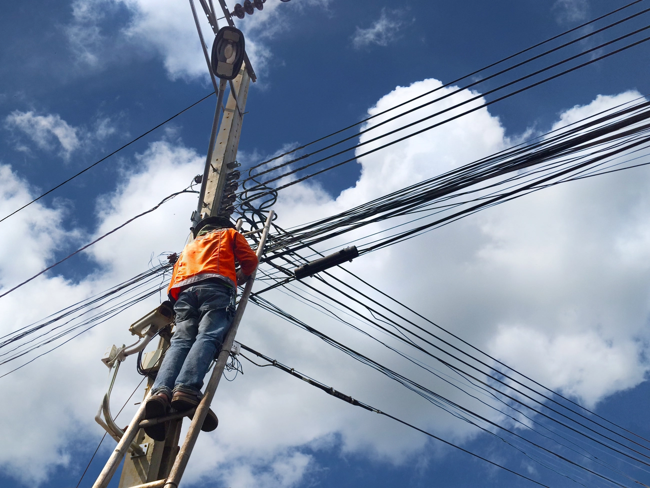 M-J tief.und Rohrleitungsbau GmbH - Der Techniker auf einer Holzleiter überprüft Glasfaserkabel an einem Strommast am Arbeitsplatz mit unordentlichen elektrischen Kabeln an einem Laternenpfahl vor blauem Himmelshintergrund.